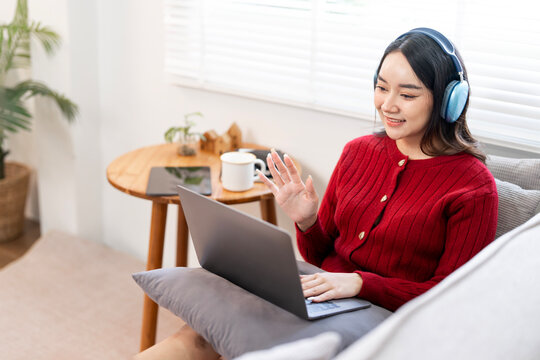 Smiling young Asian woman wearing headphones waving while engaged in a video call or online class on her laptop