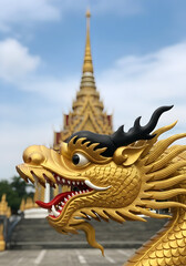 Fototapeta premium Majestic golden dragon head sculpture in sharp focus with a blurred traditional Thai Buddhist temple and its towering spire against a bright azure sky depicting spiritual heritage and cultural beauty
