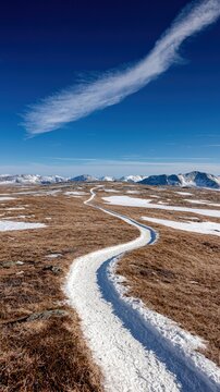 A winding, snow-covered path curves through a landscape of dry, brown grass and sparse snow patches, leading towards distant, snow-capped mountains under a vibr