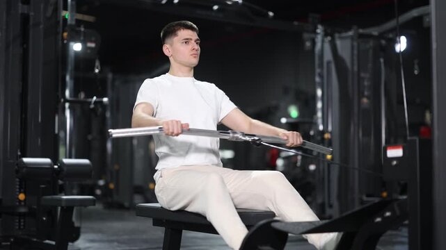 In gym, guy uses cable row machine to strengthen arm muscles. Series of repetitions to consolidate result. Perseverance and persistence in achieving fitness goal.