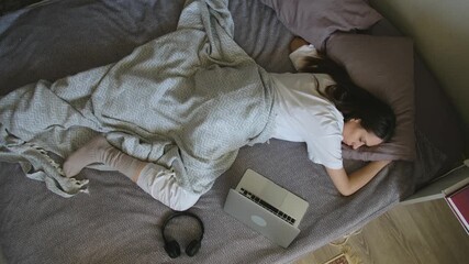Exhausted young woman napping on her bed in the middle of the day, with a laptop and headphones nearby, representing burnout, overwork, fatigue, and the need for rest and relaxation