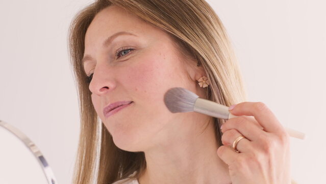 Feminine visage. Natural makeup. Attractive confident female using brush to apply powder on face skin looking in mirror on white.