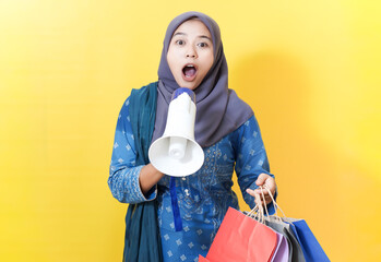 Excited woman in hijab with megaphone and shopping bags on yellow background.