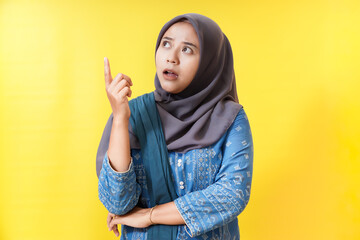 Woman in hijab pointing up with surprised expression on yellow background.
