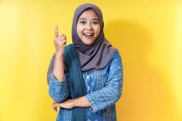 Excited woman in hijab pointing up on yellow background.