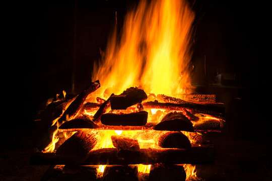 Narrow focus of a crackling bonfire at night in Brazil. Orange flames dance among stacked wood, illuminating the darkness. Sparks fly up, creating a fascinating visual spectacle.