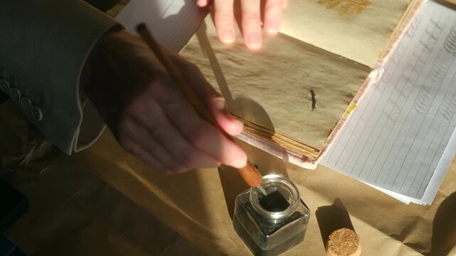 Close up calligrapher hands dipping classic nib pen into an inkwell and writing in vintage journal, practicing art beautiful handwriting on aged paper in sunlight. Practicing old calligraphy nib pen