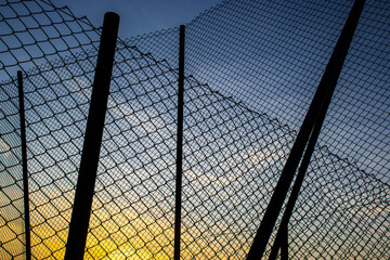 Sunset seen through a chain-link fence, the mesh silhouetted against warm sky—evoking themes of safety, boundaries and urban-nature contrast.