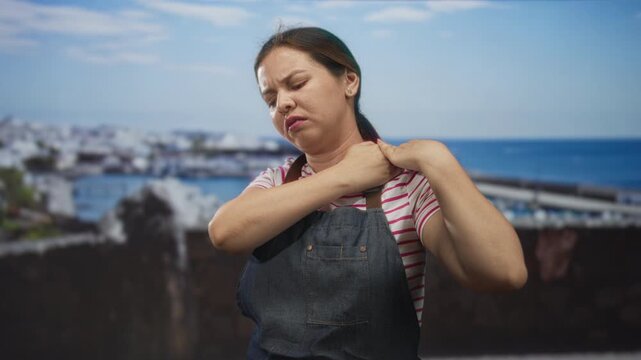 Young caucasian woman holding shoulder at seaside building wearing apron and striped tshirt near boats and harbor; pain exhaustion.