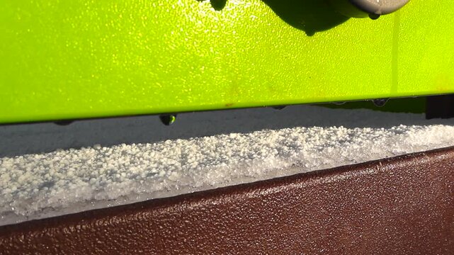 Close up view gliding in front of a playground wall where white fresh snow is melting and water drops are visible in the warm sunlight. Green and brown structure walls outside.