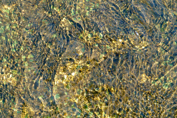 Sunlight refracts through clear, shallow water, illuminating a textured riverbed of pebbles and sand. Ripples create dynamic patterns of light and shadow, a natural abstract background.