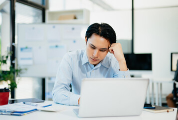 Obraz premium Asian man feeling migraine head strain.Overworked businessman financier while working on laptop and tablet at office.