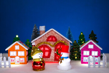 In the foreground of a small Christmas village with colorful houses there are two snowmen