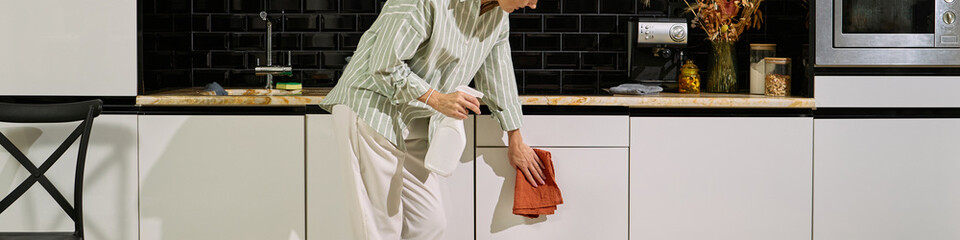 Header of Senior Caucasian woman cleaning kitchen cabinet with spray bottle and cloth, standing in modern kitchen, focusing on household chores, partially visible upper body