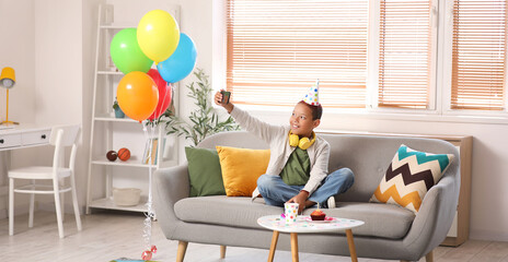 Cute little African-American boy in party hat celebrating birthday and taking selfie on sofa in living room