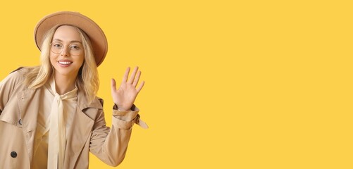 Beautiful young stylish woman waving hand on yellow background with space for text