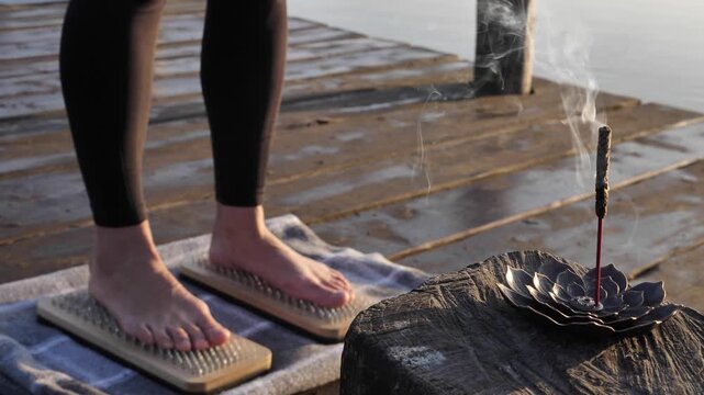 A woman stands barefoot on Sadhu boards (board of nails) on a wooden deck near the water, engaging in a meditation or extreme wellness practice next to a burning incense stick