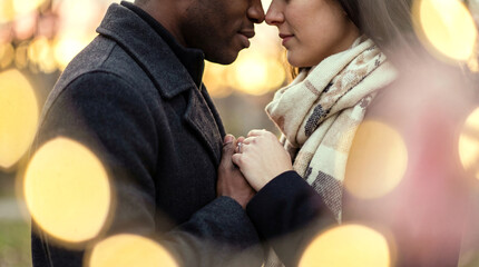 Young diverse couple holding hands and embracing with faces touching, surrounded by warm bokeh lights outdoors during winter