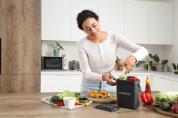 Young African-American woman throwing vegetables into container for compost on table in kitchen