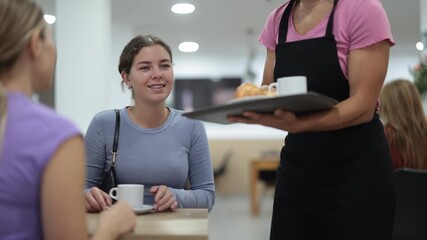 Adult female waiter serves fresh coffee and croissant to female customers in cafe