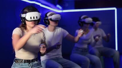 In darkened hall of gaming club, girl in VR helmet takes part in group game. Player overcomes obstacles and reaches goal in parallel reality. Imaginary fantastic world 
