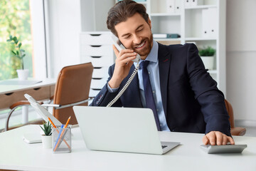 Fototapeta premium Young male secretary with laptop and calculator talking by telephone at table in office
