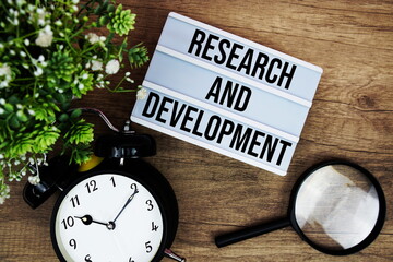 Research And Development text in lightbox with magnifying glass and alarm clock flat lay on wooden background