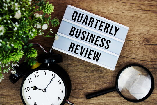 QBR Quarterly Business Review text in lightbox with magnifying glass and alarm clock flat lay on wooden background