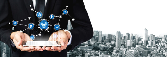 A businessman in a suit stands holding a tablet displaying various digital icons, symbolizing technology and innovation against a modern urban landscape background. Tessel