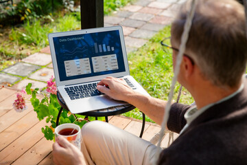Middle-aged man working remotely from laptop and drinking coffee in hanging chair on sunny patio, relaxed lifestyle and modern home office.