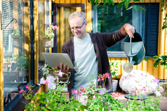 Middle-aged man watering flowers and working remotely on laptop during video call on sunny patio, modern multitasking lifestyle concept.