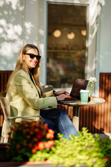 Woman working on laptop at an outdoor cafe surrounded by plants and sunlight