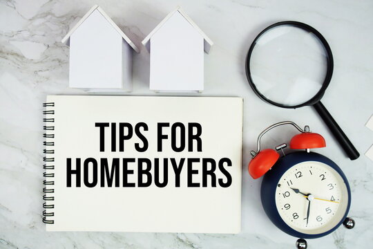 Tips For Homebuyers text written on notepad with house model, magnifying glass and alarm clock on marble background