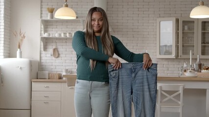 Cheerful attractive plus size African American woman in casual clothes showing oversized jeans, celebrating successful weight loss diet plan, expressing happiness and excitement at home.