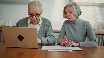 Old woman and her husband counting income and expenses at home kitchen, portrait . Senior lady viewing utility bills and aged man checking it online in laptop, distant payment service on bank web-site