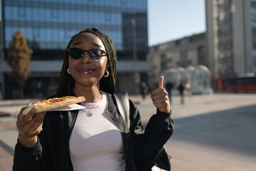Beautiful black woman with sunglasses eating a slice of pizza on the city street on a sunny day