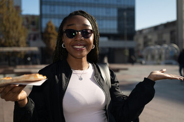 Beautiful black woman with sunglasses eating a slice of pizza on the city street on a sunny day