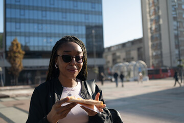 Beautiful black woman with sunglasses eating a slice of pizza on the city street on a sunny day