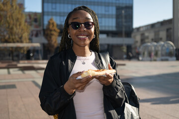 Beautiful black woman with sunglasses eating a slice of pizza on the city street