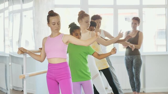 Group of concentrated teenage students of ballet school diligently practicing Demi rond de jambe par terre at barre in sunlit mirrored choreographic studio to applause of smiling female choreographer