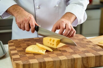 Professional chef hands cutting block of yellow cheese with holes using knife on wooden board. Cook in white uniform slicing dairy food in restaurant kitchen. Food preparation process closeup.