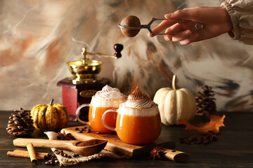 Female hand sprinkling cinnamon powder on glass cup of orange pumpkin latte with whipped cream against beige background