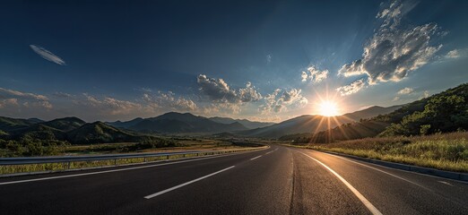 An empty asphalt road stretches towards a mountain range as the sun sets in a beautiful sky