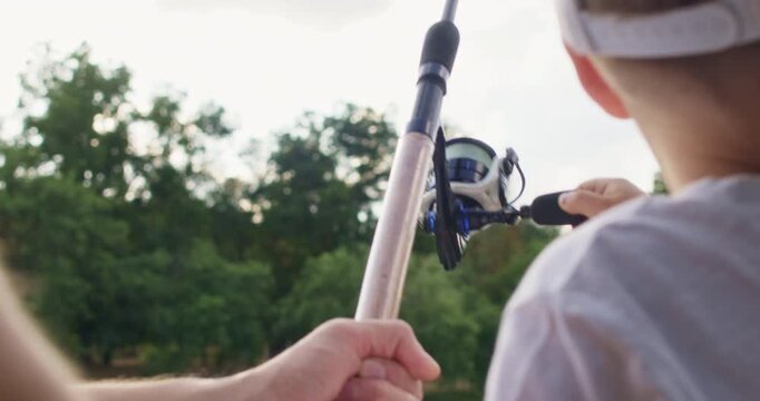 Concentrated little preschooler boy and father turn reel on fishing rod to pull fish out of water. Loving parent supports fishing rod and gives instructions