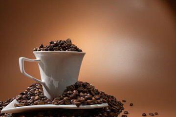 White coffee cup filled to the brim with glossy roasted beans on a matching saucer. Warm brown background and soft light create a cozy cafe atmosphere.