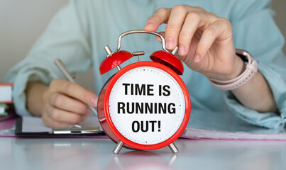 A person is writing on a piece of paper with a pen. A red alarm clock is on the table with the words Time is Running Out written on it