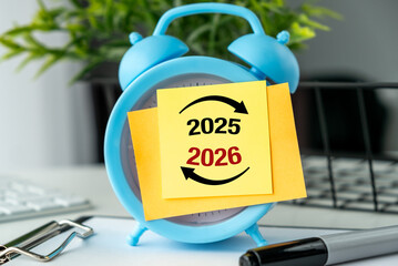 A blue alarm clock with a yellow post it says 2025 and 2026 on it. The clock is on a table with a basket and a pen