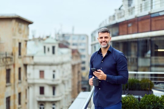Confident Professional On a City Rooftop Balcony Using Smartphone and Coffee Mug in Morning Light