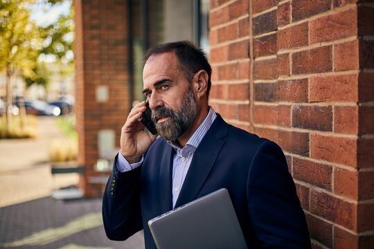 Professional Man in Navy Suit On Phone Near Brick Wall Holding Laptop Outside Office - Powered by Adobe
