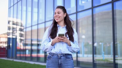 Young professional woman happily texting on her mobile phone. She is standing in front of a modern glass office building on a sunny day